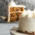 The best Carrot Cake recipe on a marble cake stand with a piece being lifted to serve.