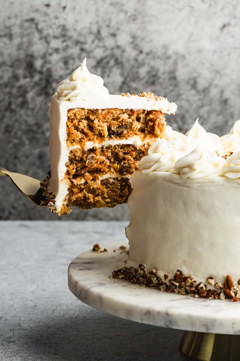 The best Carrot Cake recipe on a marble cake stand with a piece being lifted to serve.