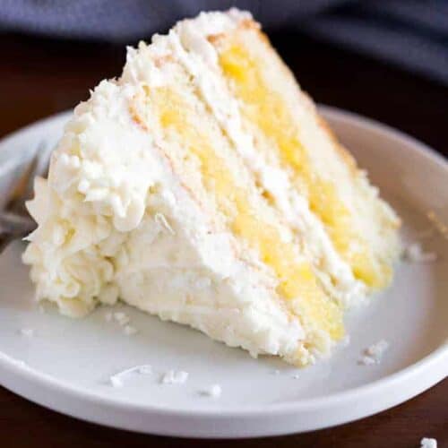 A slice of Coconut Cake on a plate, with layers of pineapple filling and frosted with coconut cream cheese frosting.