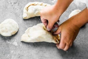 Hands sealing and crimping the edges of a Cornish pasty dough.