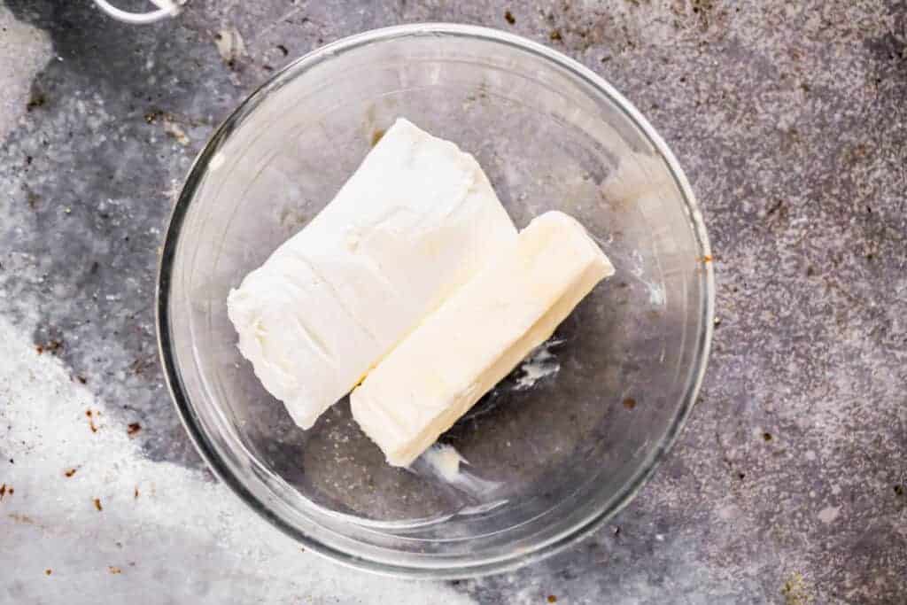 Un bâton de beurre et un bloc de fromage à la crème ajoutés dans un bol à mélanger.