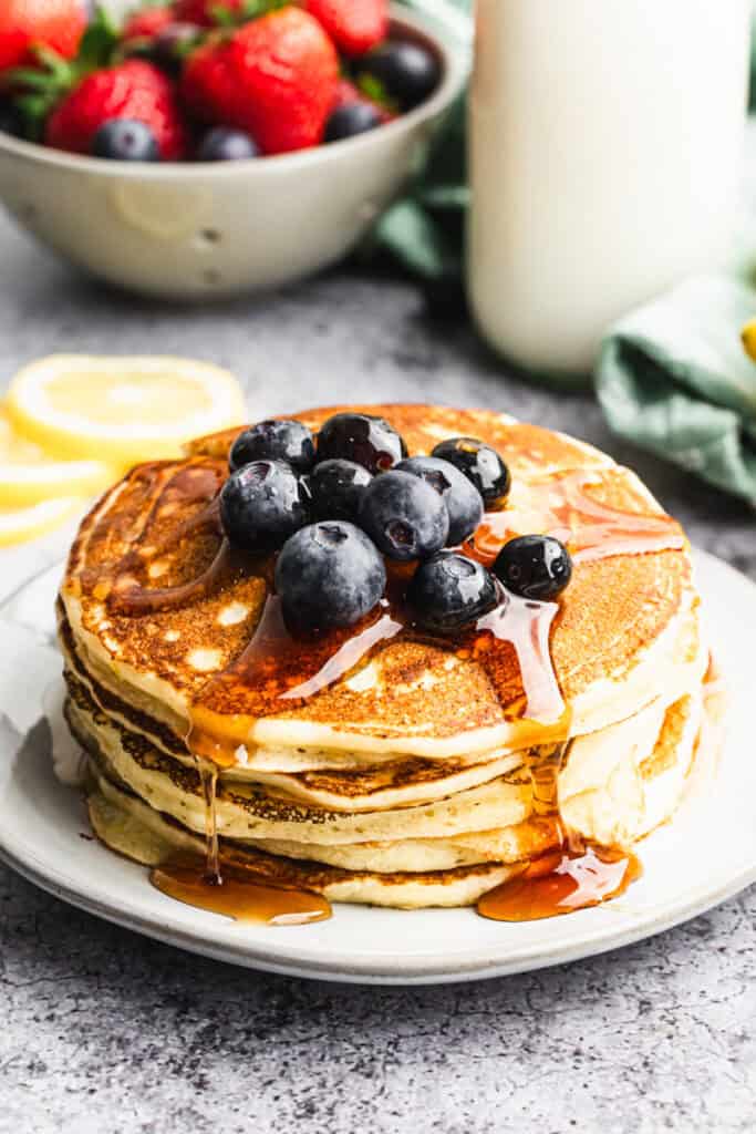 A stack of simple Lemon Ricotta Pancakes topped with fresh blueberries and drizzled with maple syrup.