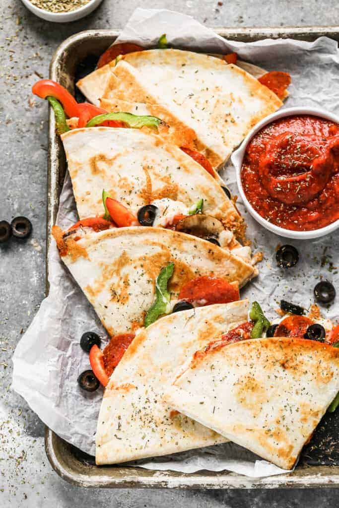 Pizza quesadillas on a baking sheet with a side of marinara sauce.