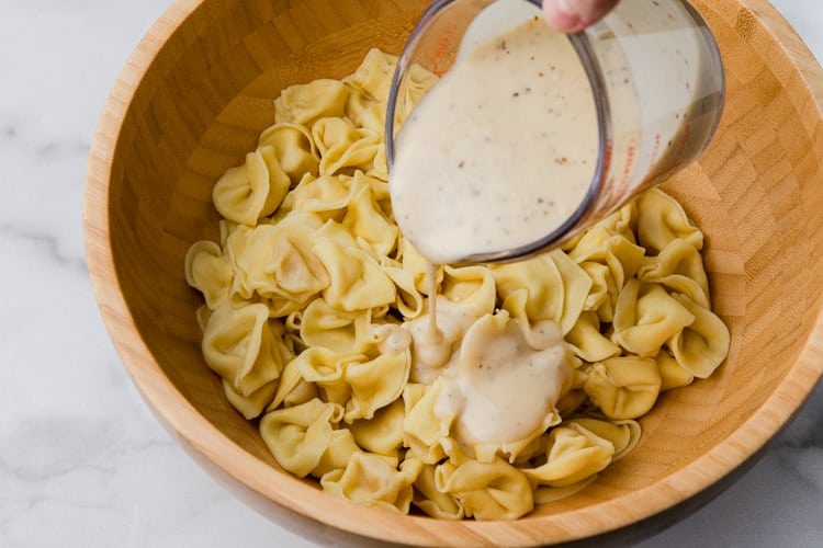 Cooked cheese tortellini in a bowl with caesar dressing being poured over them.