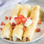 Three crepes on a plate rolled up with strawberries and cream filling, and fresh sliced strawberries and whipped cream on top.