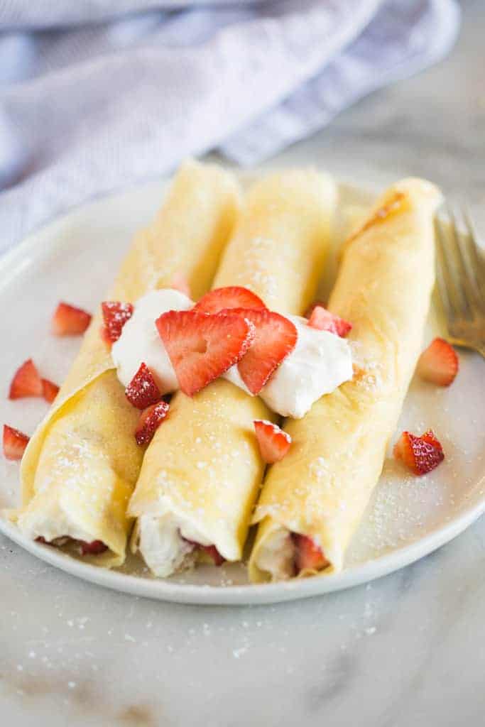 Three crepes on a plate rolled up with strawberries and cream filling, and fresh sliced strawberries and whipped cream on top.