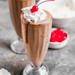 Two chocolate milkshakes in tall cups with whipped cream and cherry on top.