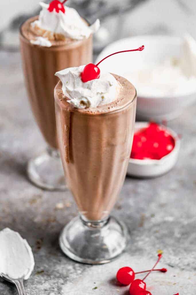 Two chocolate milkshakes in tall cups with whipped cream and cherry on top.