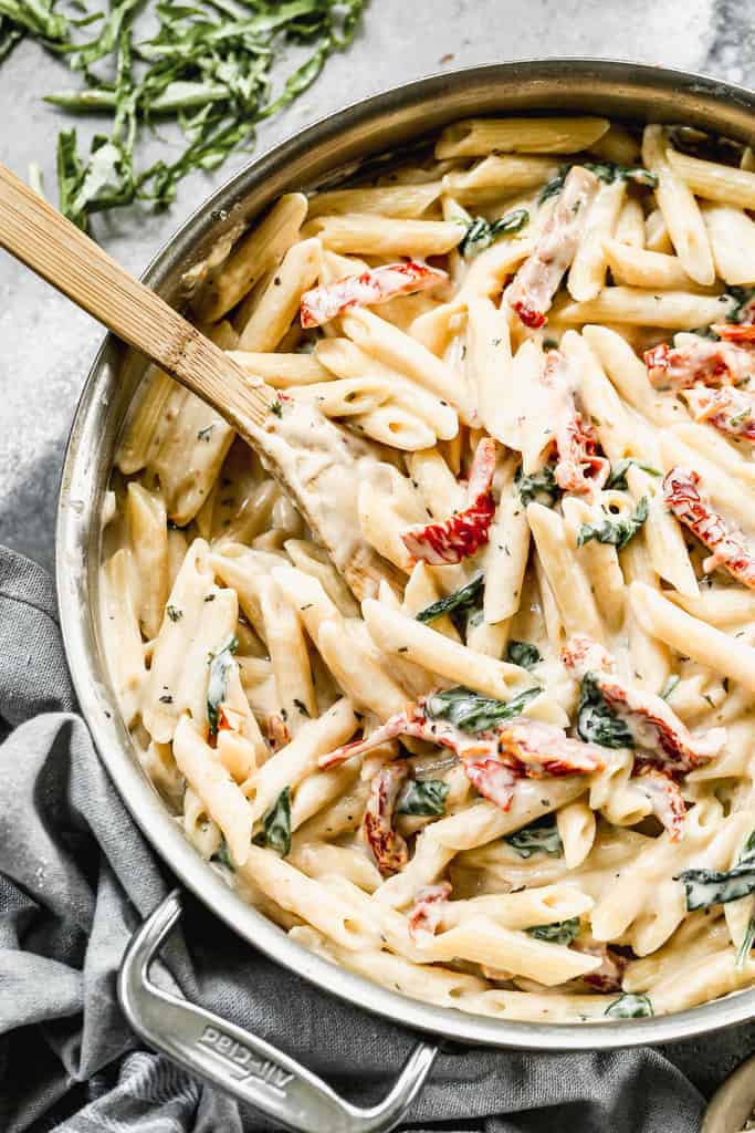 Penne pasta added to white sauce and sun dried tomatoes in a skillet.