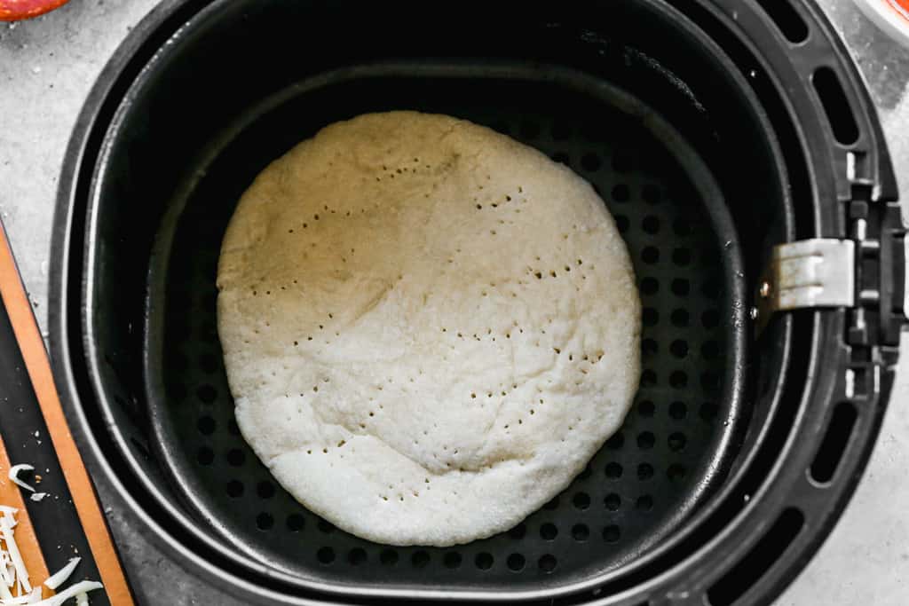 Pizza dough rolled out, pricked with a fork, and placed in an air fryer basket.