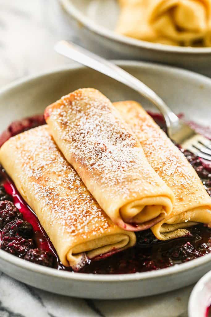Three filled blintzes stacked on a plate, with powdered sugar on top, and some berry sauce.
