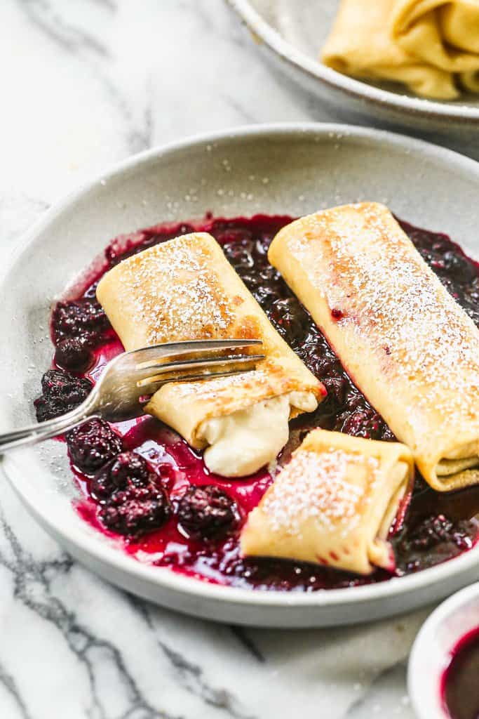 Two cheese blintzes served on a plate with berry sauce, and a fork taking a bite.