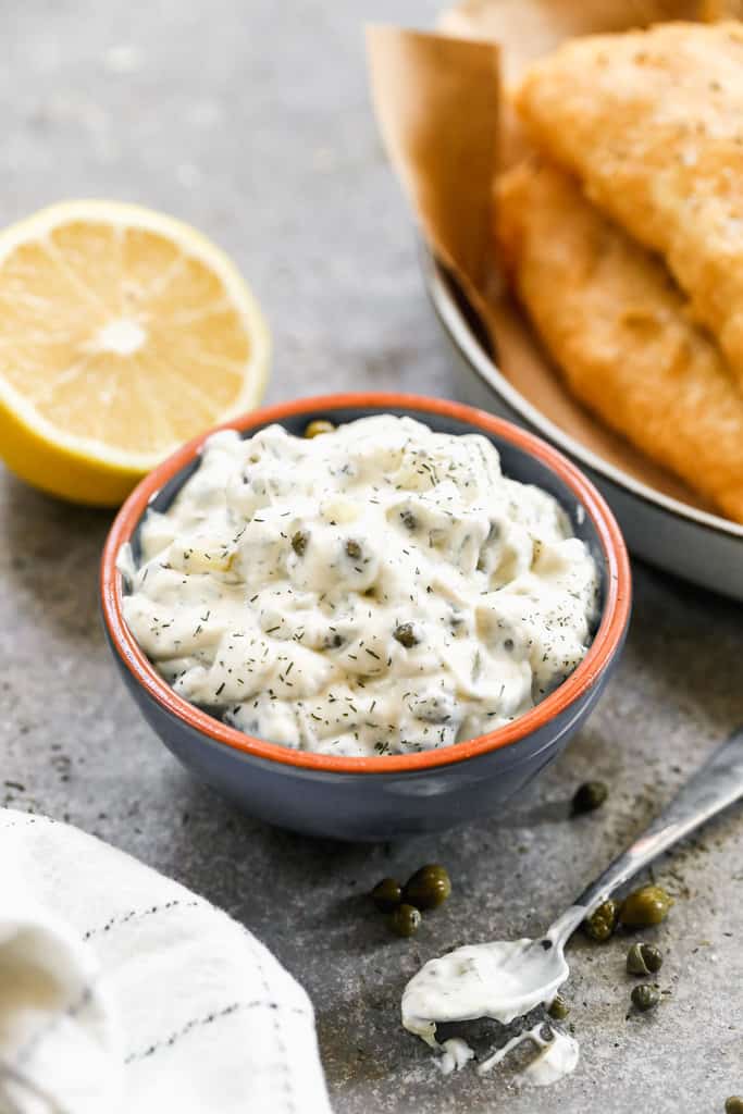 Homemade tartar sauce in a bowl next to a lemon and some fish and chips.