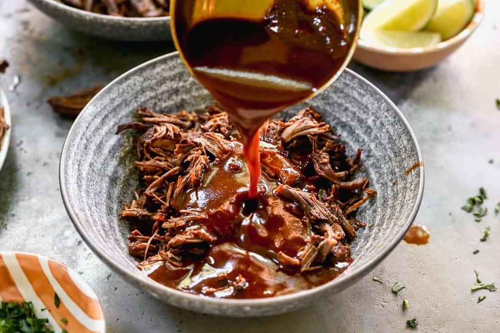 Shredded meat in a bowl with a ladle pouring birria consomé broth on top.