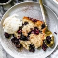 A piece of an easy Blackberry Cobbler recipe served on a plate with a scoop of vanilla ice cream and a dusting of cinnamon.