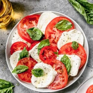 An Italian Caprese Salad recipe with slices of tomato and mozzarella arranged on a plate then topped with fresh basil, olive oil, salt, and pepper.