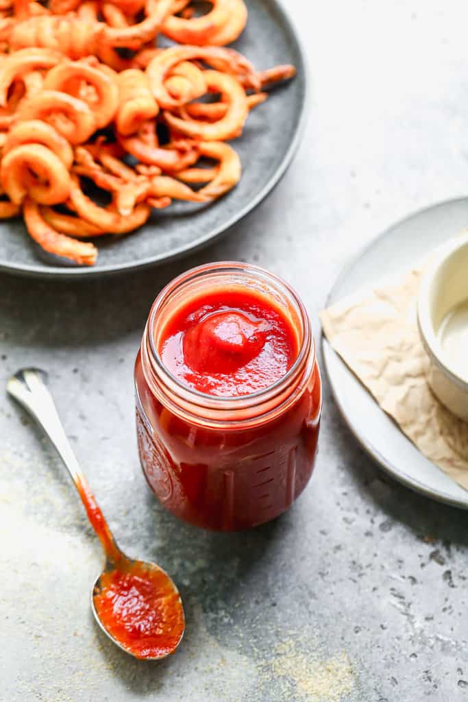 A jar of easy Homemade Ketchup with a spoon and a plate of curly fries in the background.