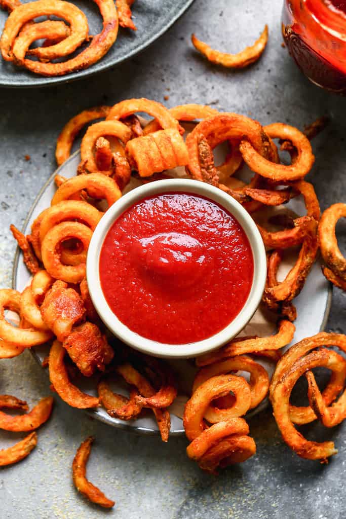 A small serving bowl of tomato ketchup on a plate surrounded by curly fries.