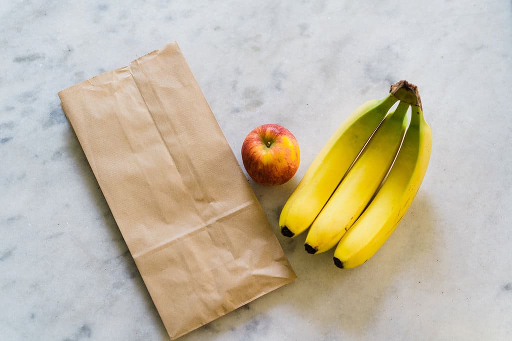 Sebuah kantong kertas cokelat, apel, dan tiga pisang bersebelahan.