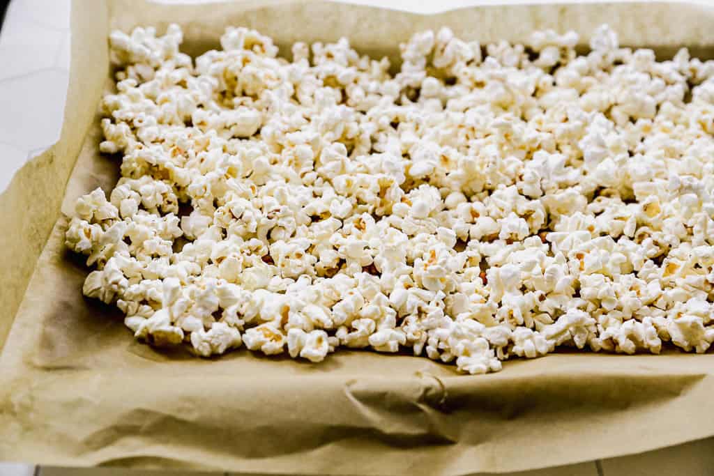 A cookie sheet lined with brown parchment paper with popcorn spread out, ready to be made into caramel popcorn.