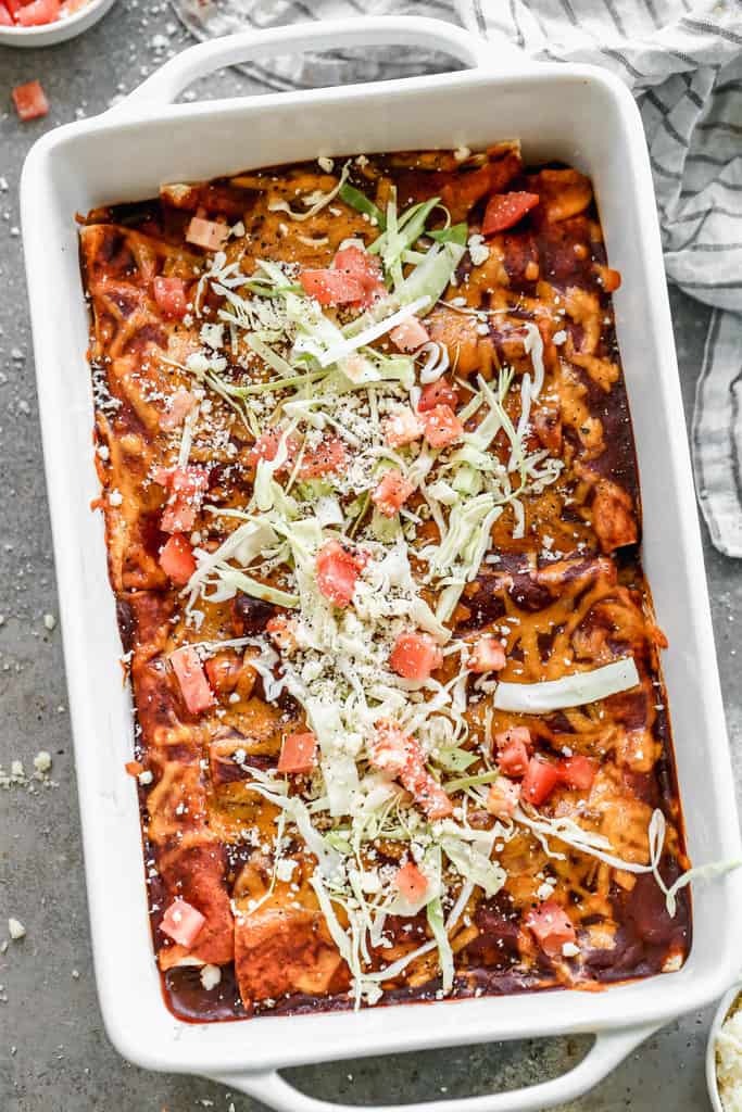 A white rectangle baking dish filled with Cheese Enchiladas and topped with shredded cabbage, cotija cheese, and tomatoes.