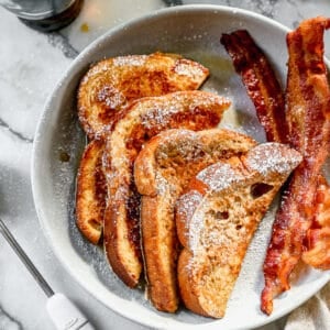 Two slices of the best French Toast cut in half and served with powdered sugar, syrup, and a side of bacon.