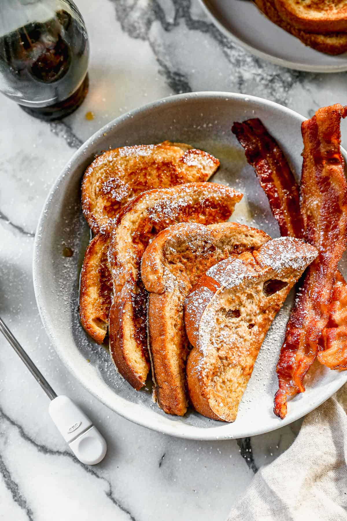 A plate with two slices of french toast cut in half with powdered sugar and syrup drizzled on top, next to two slices of bacon.