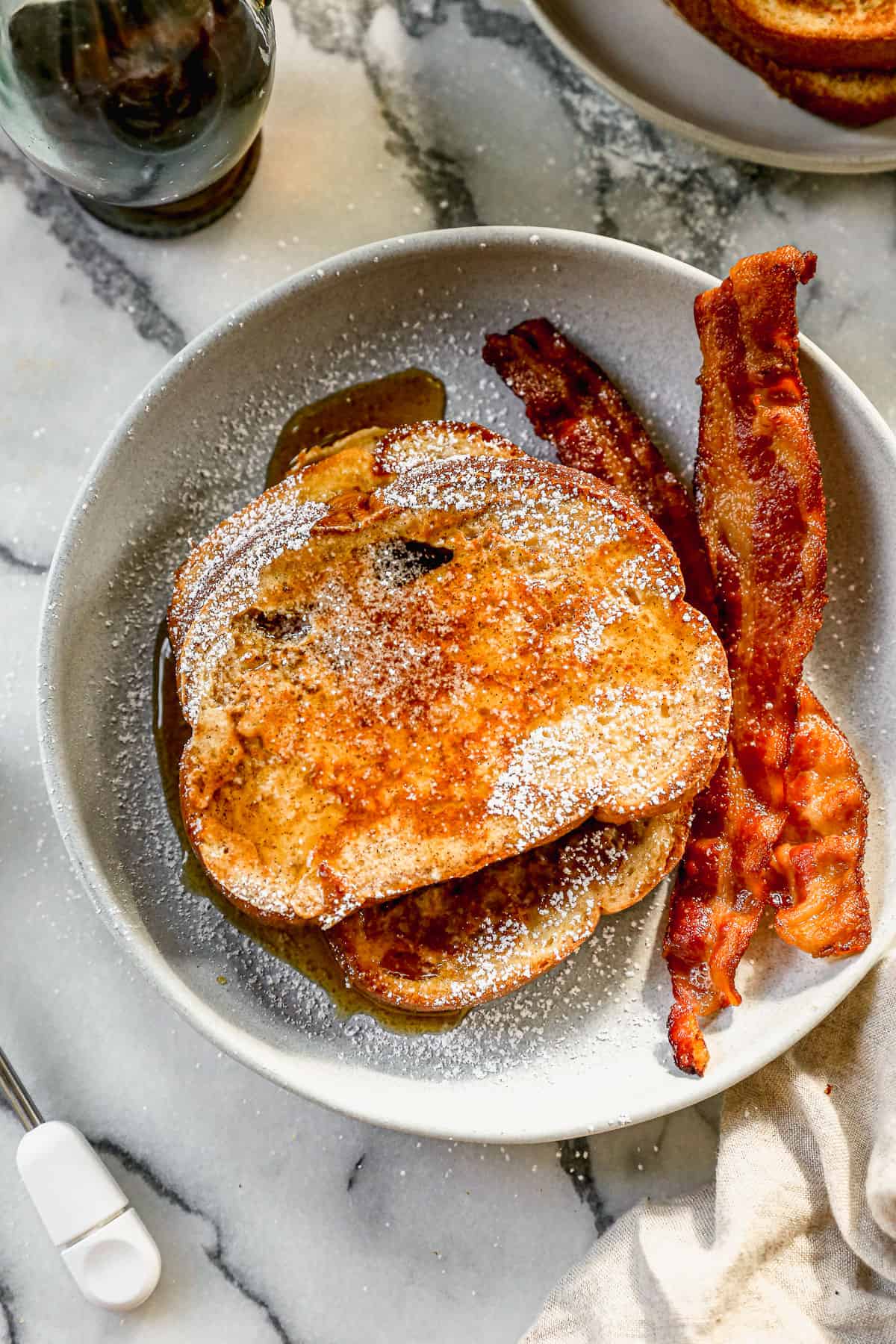Two stacked slices of the best French Toast with powdered sugar, syrup, and a side of bacon.