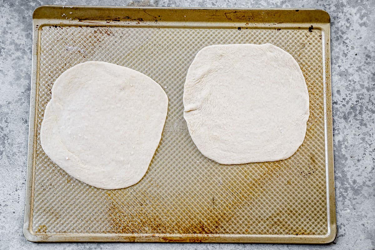 Dua kulit adonan pizza digulung dan diletakkan di atas loyang, siap untuk dipanggang terlebih dahulu dan ditambahkan topping. 