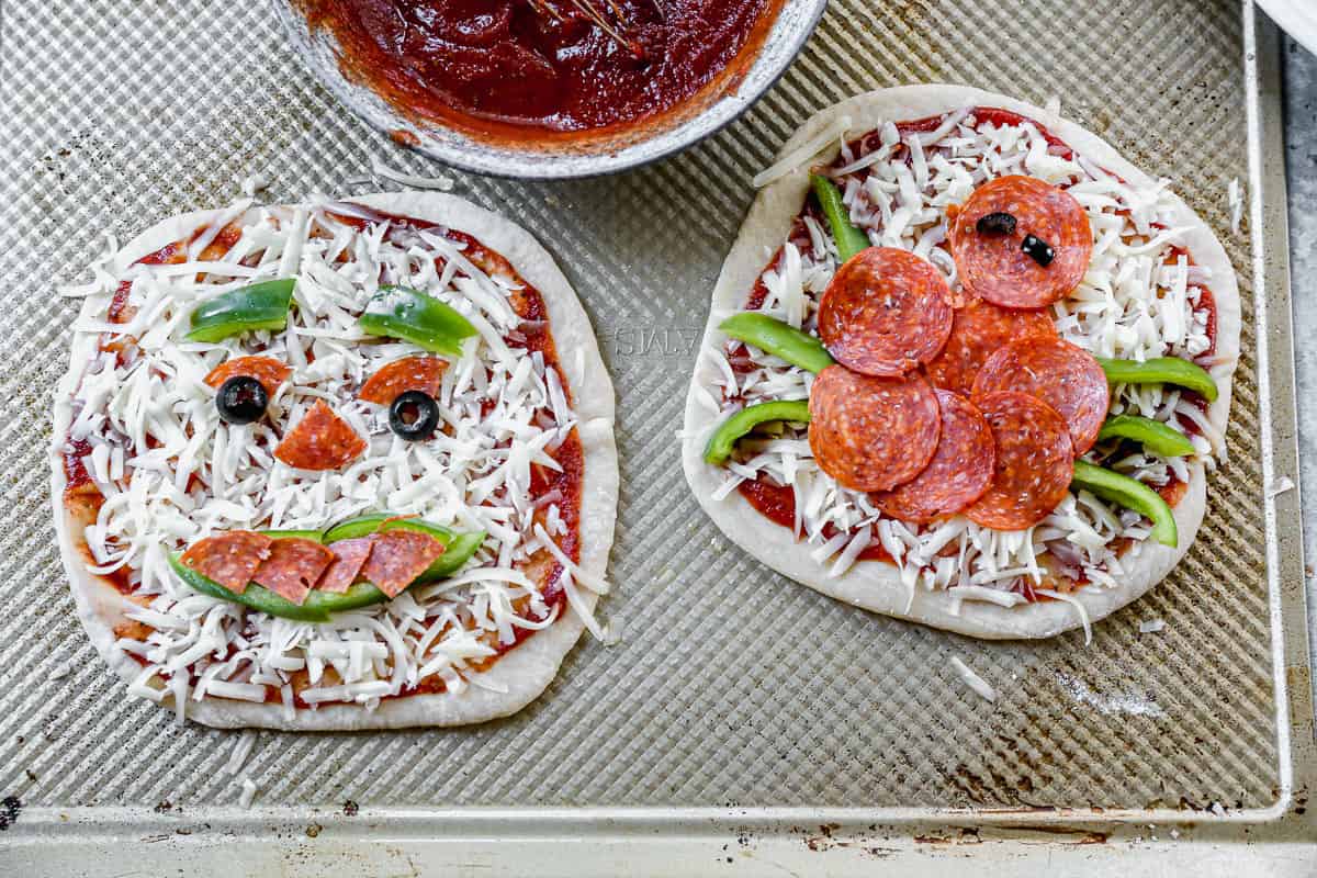 A spider pizza and a jack-o-lantern pizza on a baking sheet, ready to bake. The toppings are pepperoni, olives, and green peppers.  