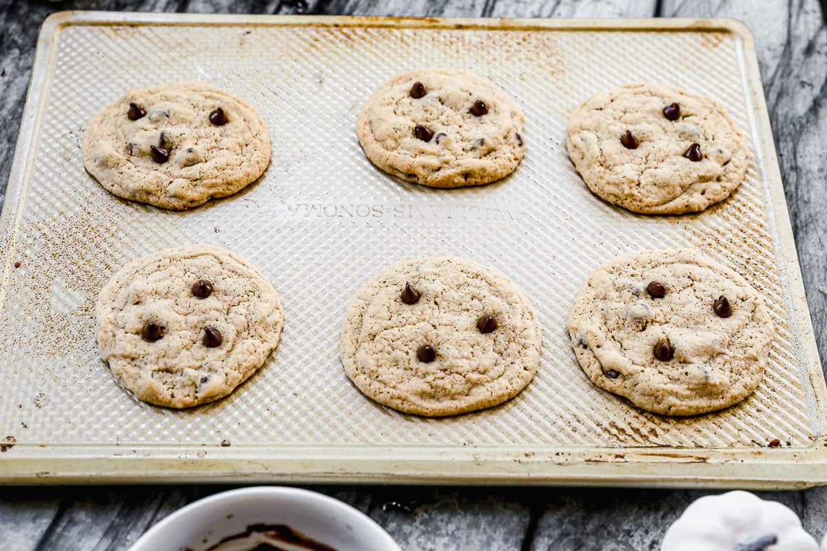 Six chocolate chip cookies on a baking sheet, with 3 chocolate chips pressed on each one. 