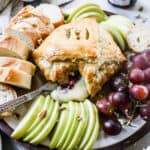 Baked Brie wrapped in puff pastry with some of the cheese coming out on a platter with apples, grapes, and bread for dipping.
