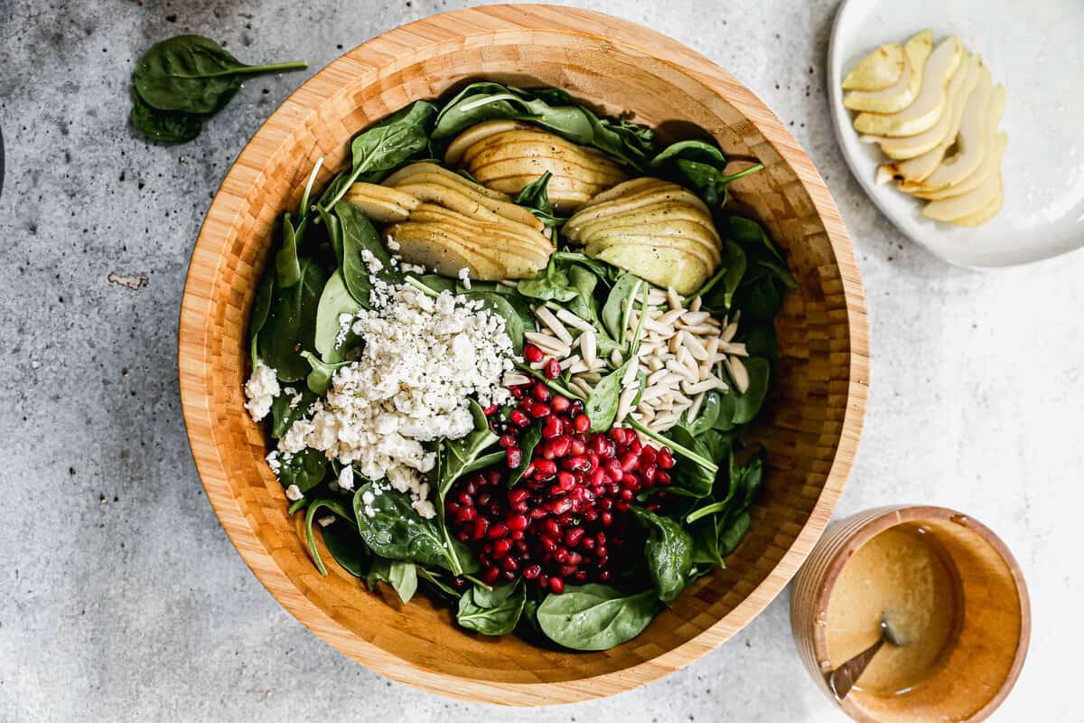A brown serving bowl with spinach, pomegranate, sliced pears, feta cheese, and slivered almonds to make a salad.