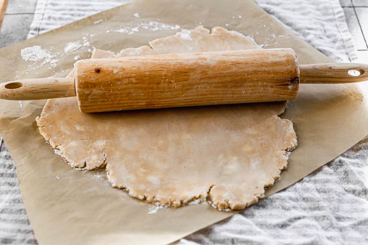 Pie dough being rolled out with a wooden rolling pin on top of parchment paper.