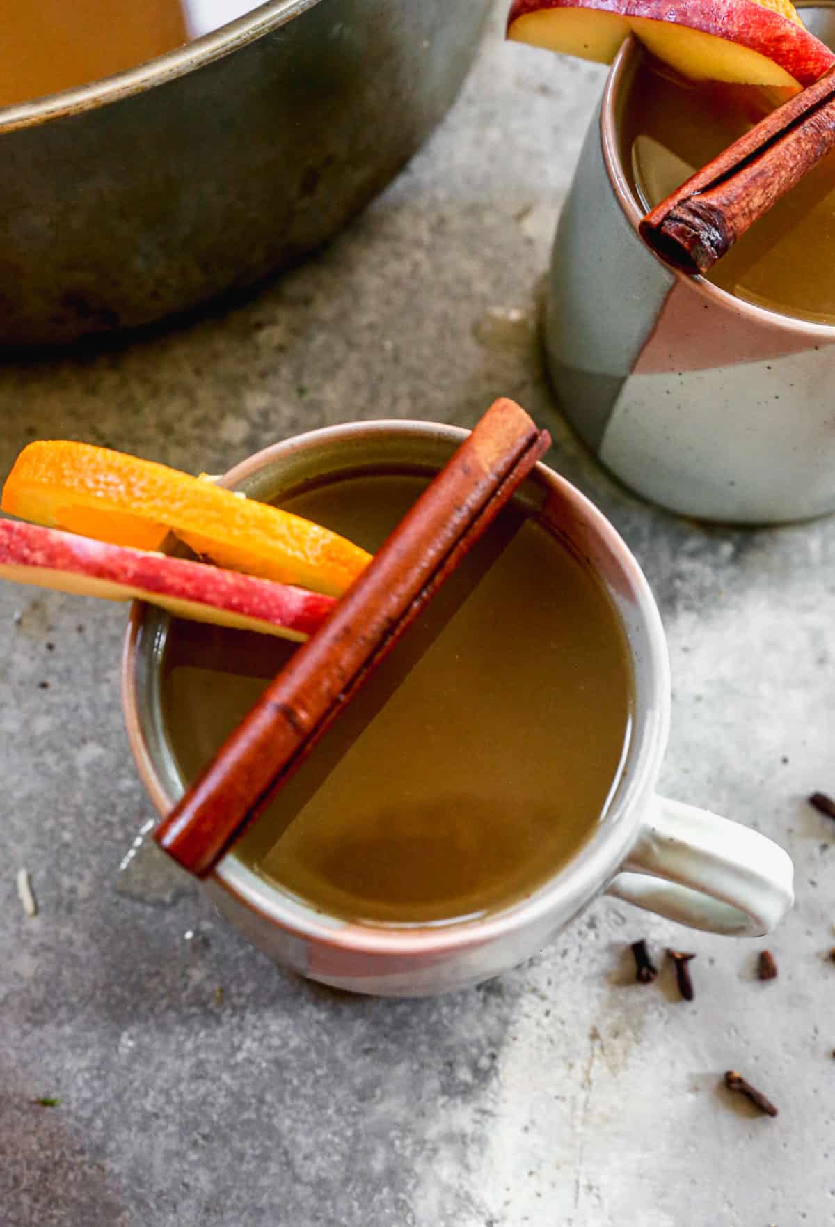 A close-up image of a mug filled with homemade Wassail and a cinnamon stick, apple slice, and orange slice on top of the mug. 