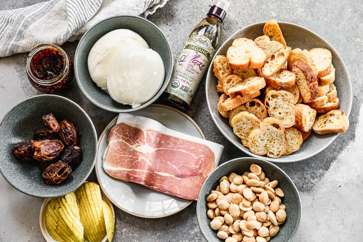 All the ingredients needed to make the best burrata appetizer: burrata, balsamic reduction, toasts, dates, preserves, prosciutto, marcona almonds, and pears.