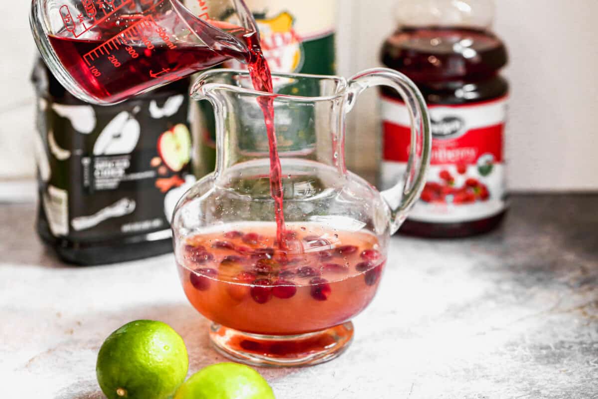 A homemade cranberry syrup in a large glass pitcher with apple cider, cranberry juice, and lime juice being added to it. 