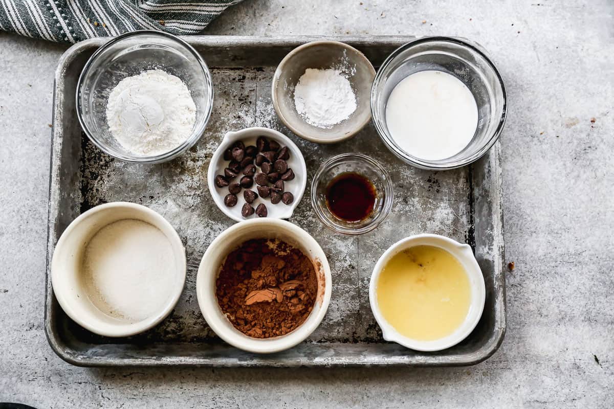 All of the ingredients needed to make the best chocolate mug cake recipe on a baking sheet: flour, sugar, cocoa powder, baking powder, salt, milk, canola oil, vanilla, and chocolate chips.