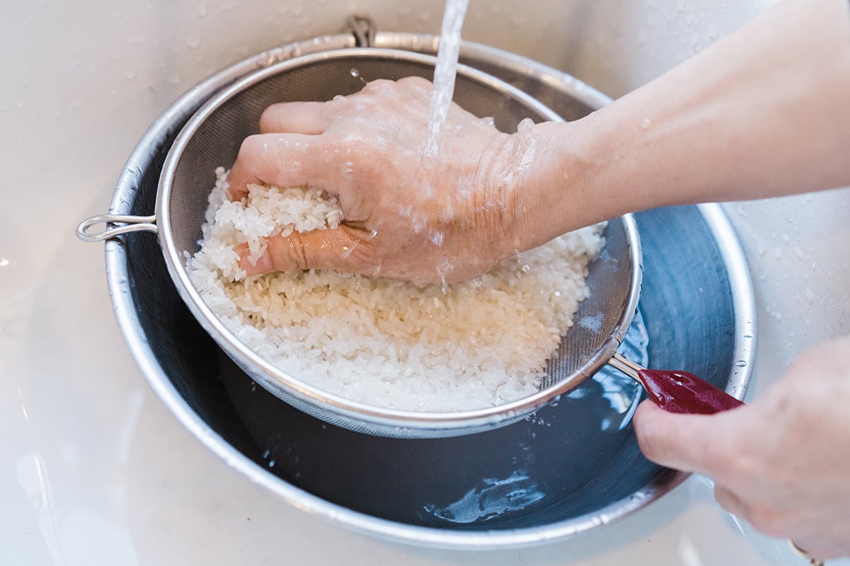 Rice for sushi being washed in a fine mesh strainer with a bowl underneath.