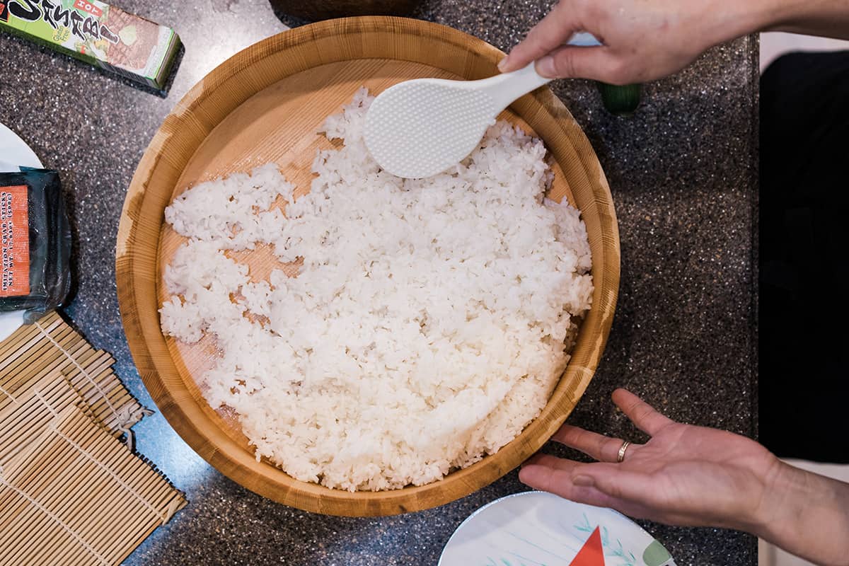 A wet wooden bowl filled with sushi rice and being sliced and cooled with a flat plastic paddle.