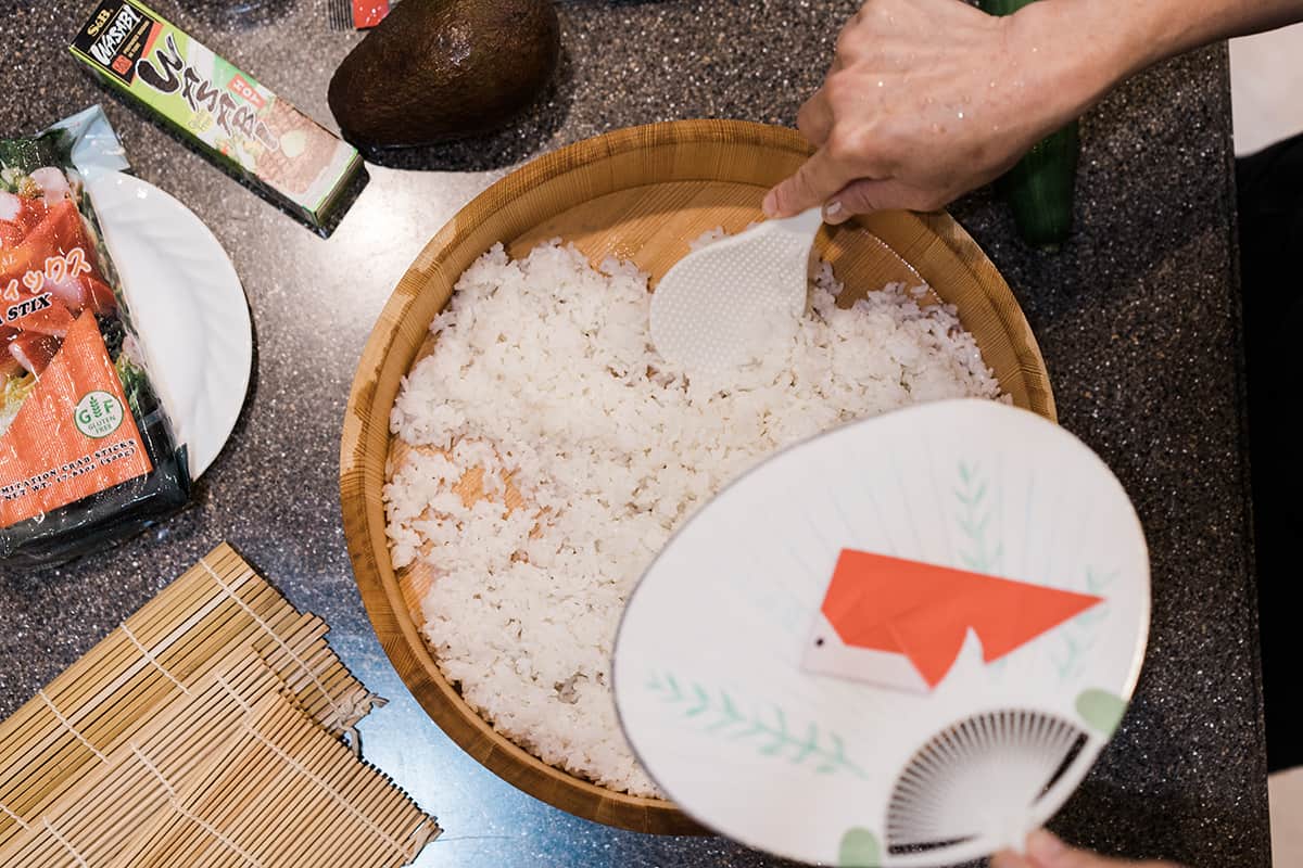 Sushi rice in a large flat wooden bowl being cooled by a Japanese fan.