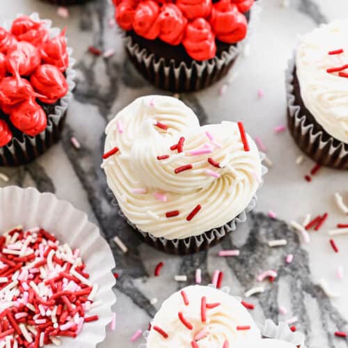 A white frosted heart cupcake with Valentine sprinkles surrounded by red and white frosted heart cupcakes.