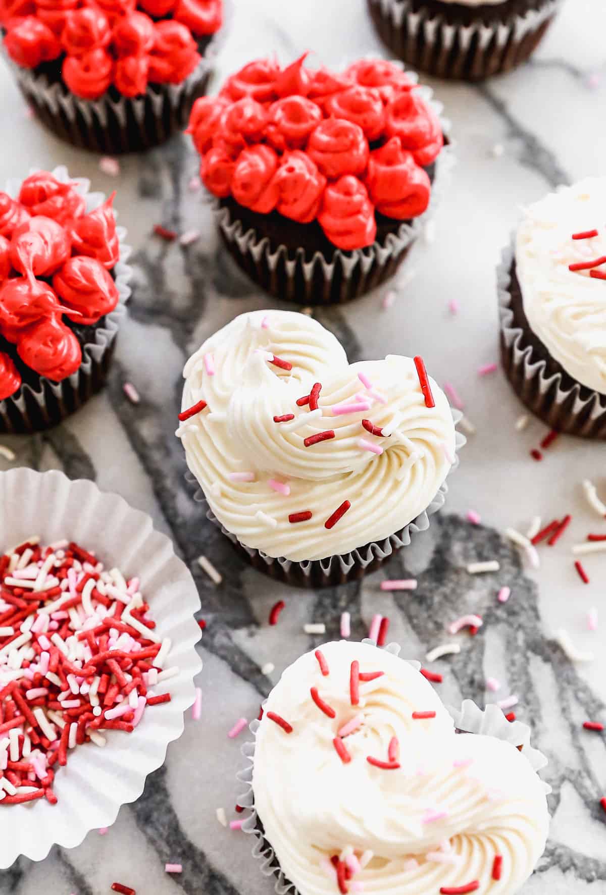 A white heart shaped cupcake with Valentine's sprinkles, surrounded by more red and white frosted heart cupcakes.