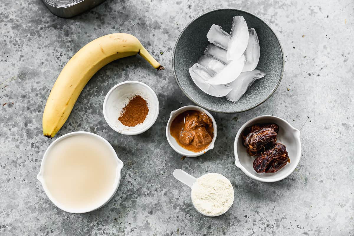 All the ingredients needed for the best vanilla protein shake: banana, cinnamon, almond butter, pitted dates, protein powder, almond milk, and ice.