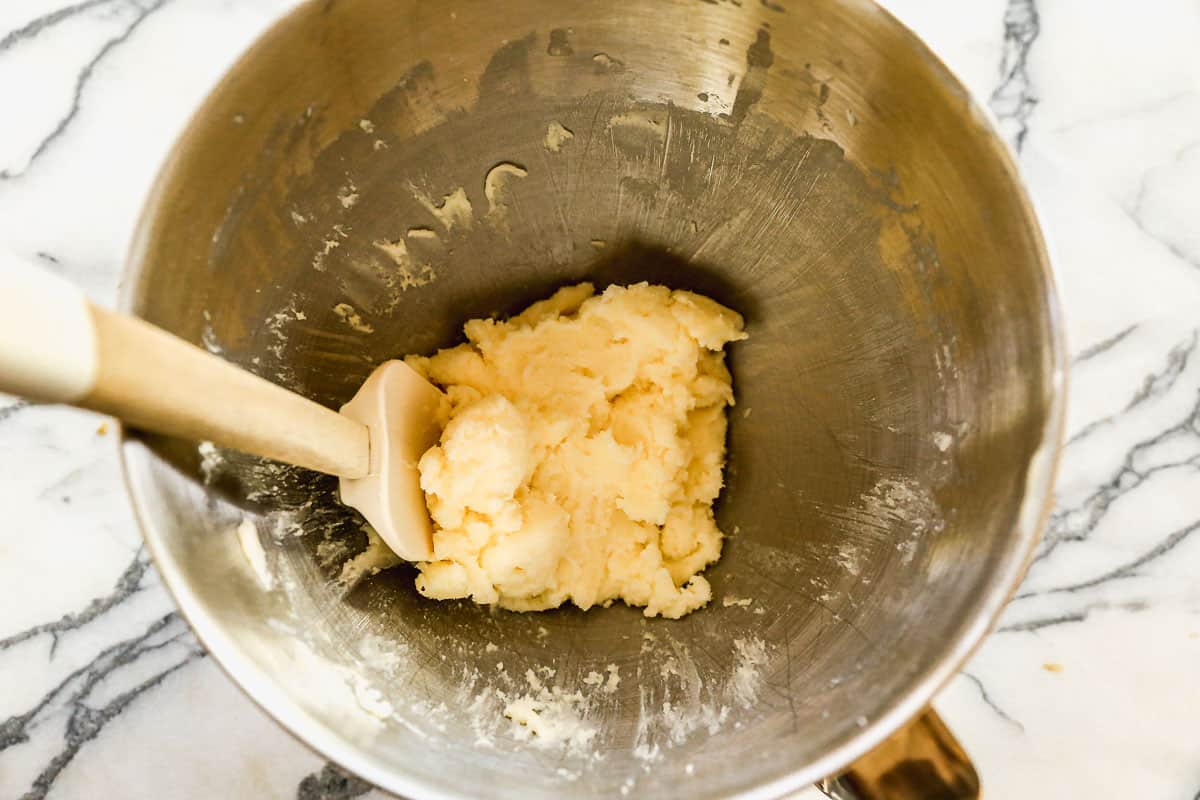A stainless steel mixing bowl with a rubber scraper and butter and sugar creamed together.