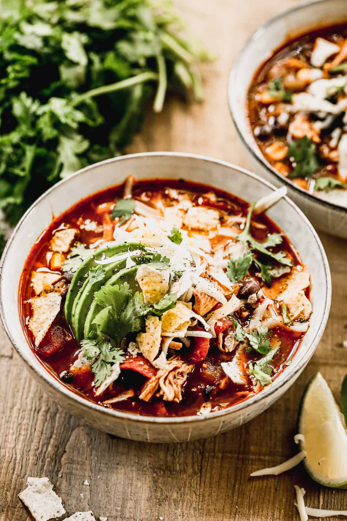 A bowl of Chicken Tortilla Soup with crumbled tortilla chips, cilantro, cheese, and sliced avocado.