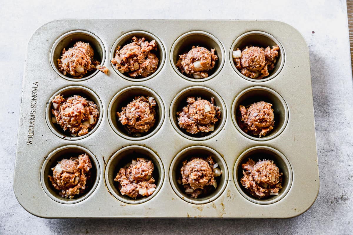 A scoop of meatloaf mixture in each compartment of a muffin pan.