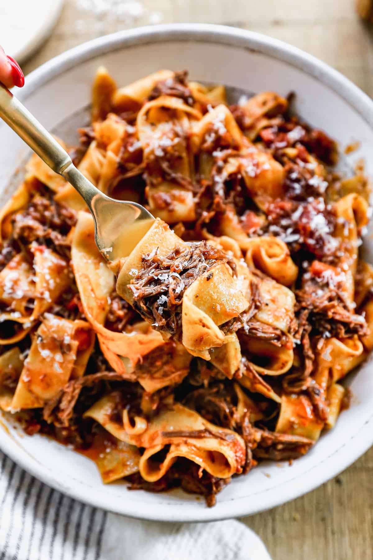 A fork lifting up a bite of the best Short Rib Ragu sprinkled with fresh parmesan cheese.