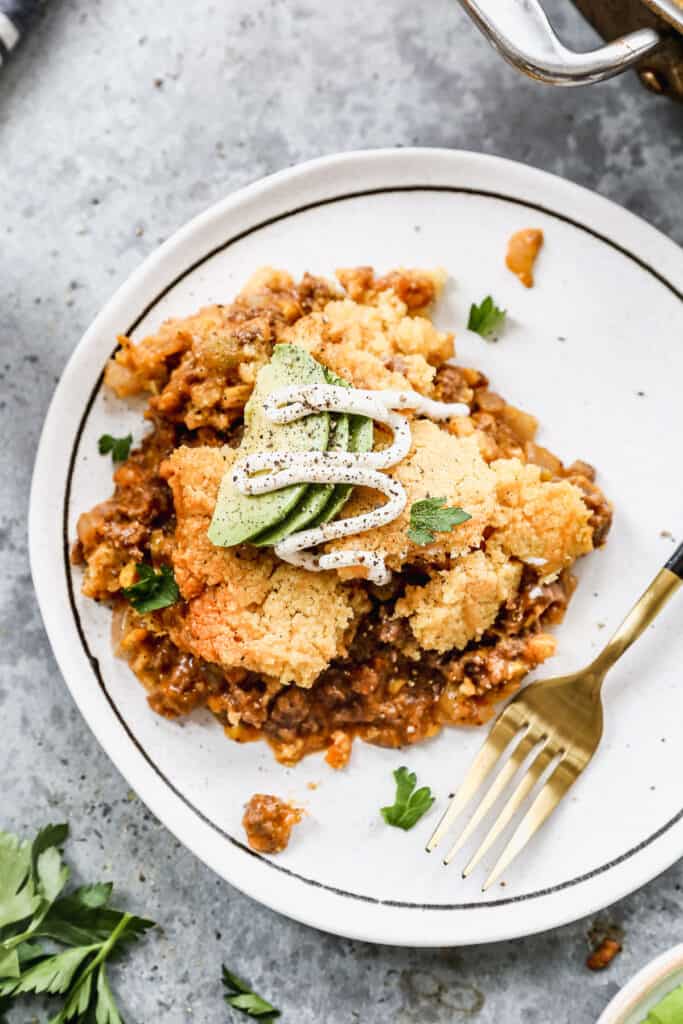 A serving of homemade Tamale Pie on a white plate, topped with fresh avocado slices, sour cream, and salt and pepper.