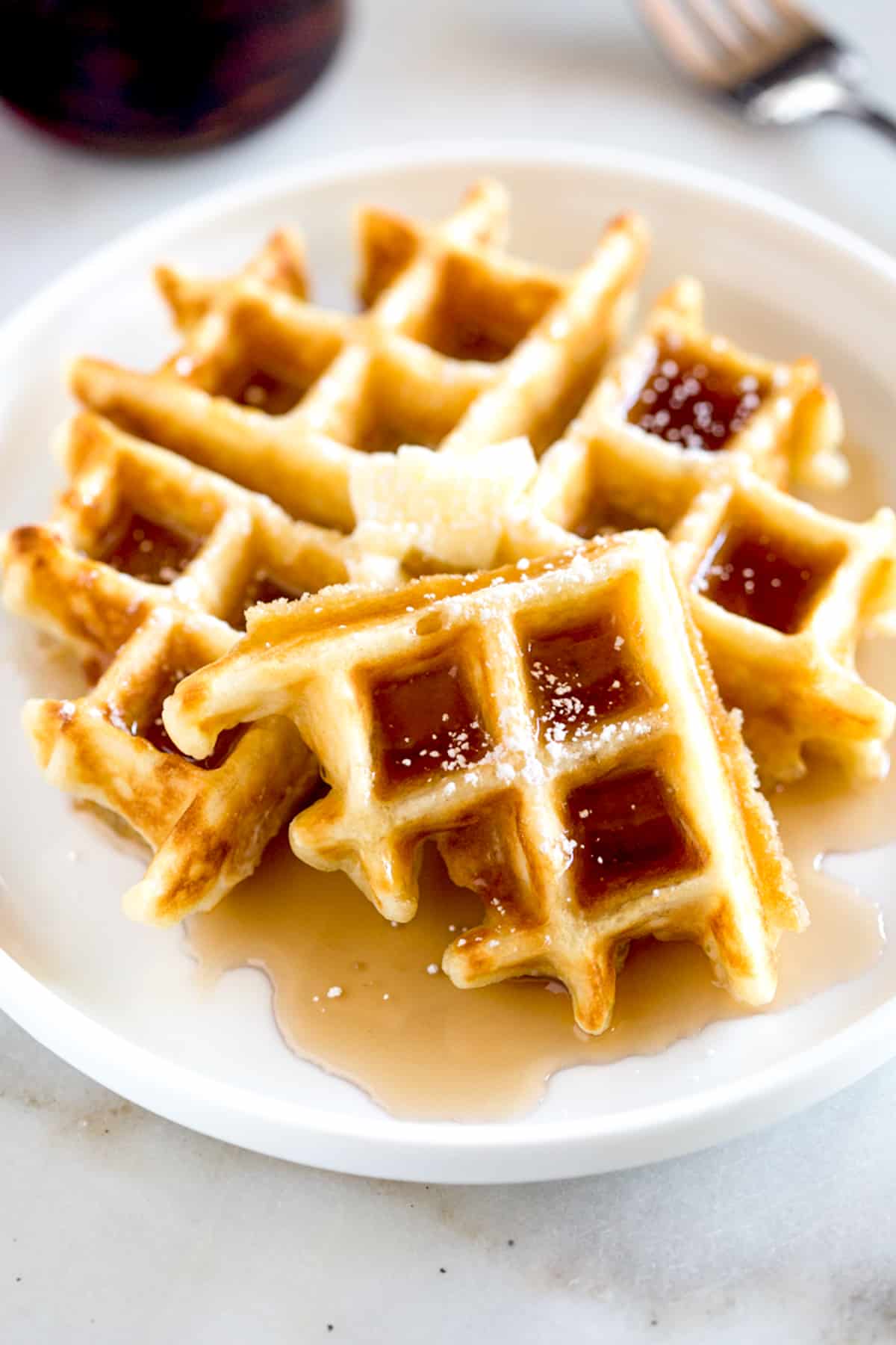 A close image of an easy Belgian waffle topped with butter, syrup, and powdered sugar; with one corner broken off and leaning against the waffle. 